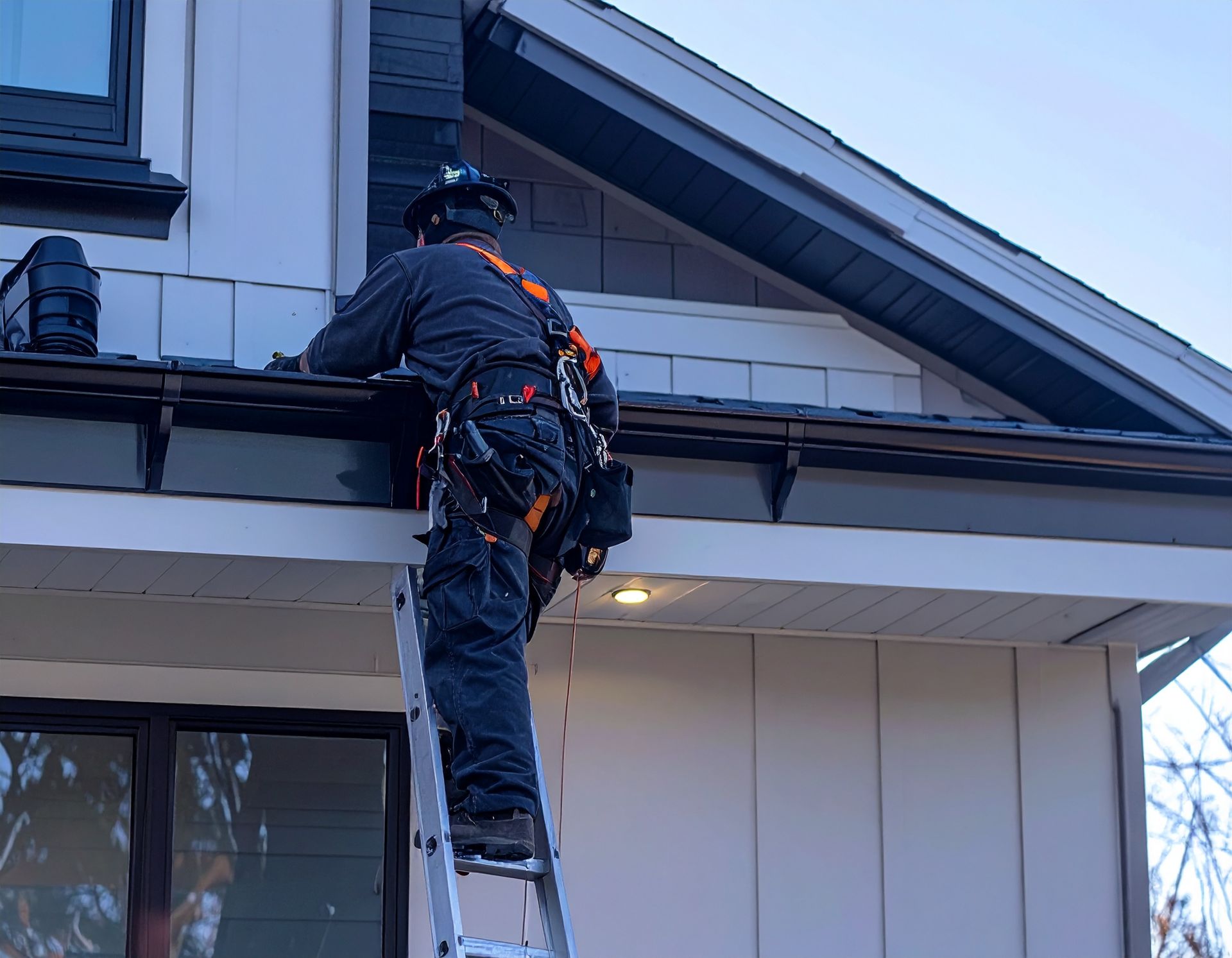 A worker in safety gear climbs a ladder to inspect a house roof and gutter.