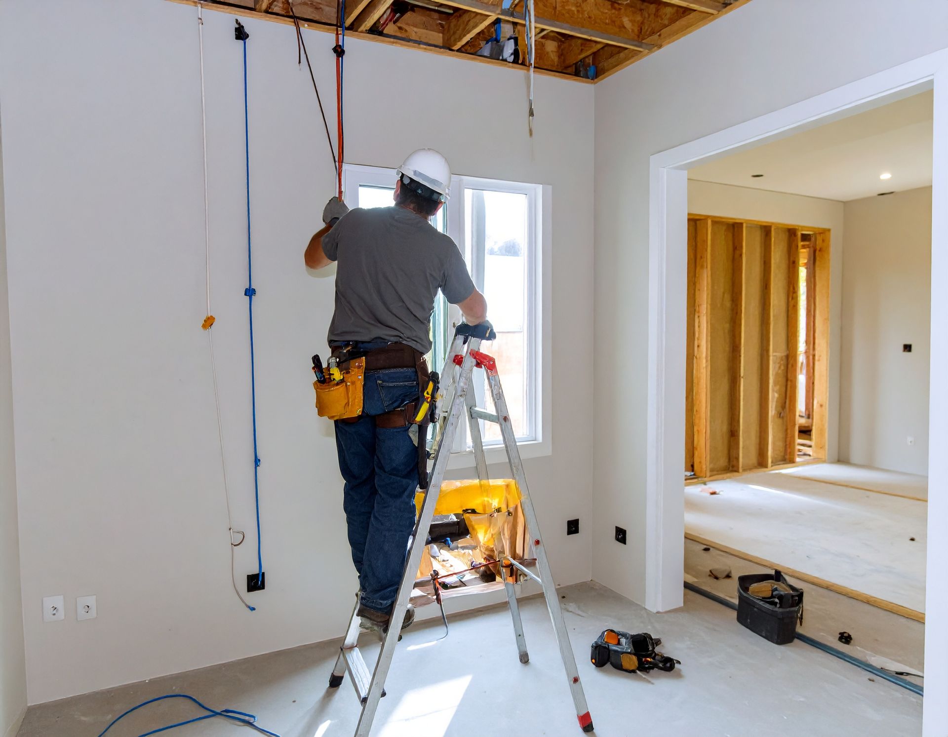 Worker on a ladder installing wiring in a white room under construction.