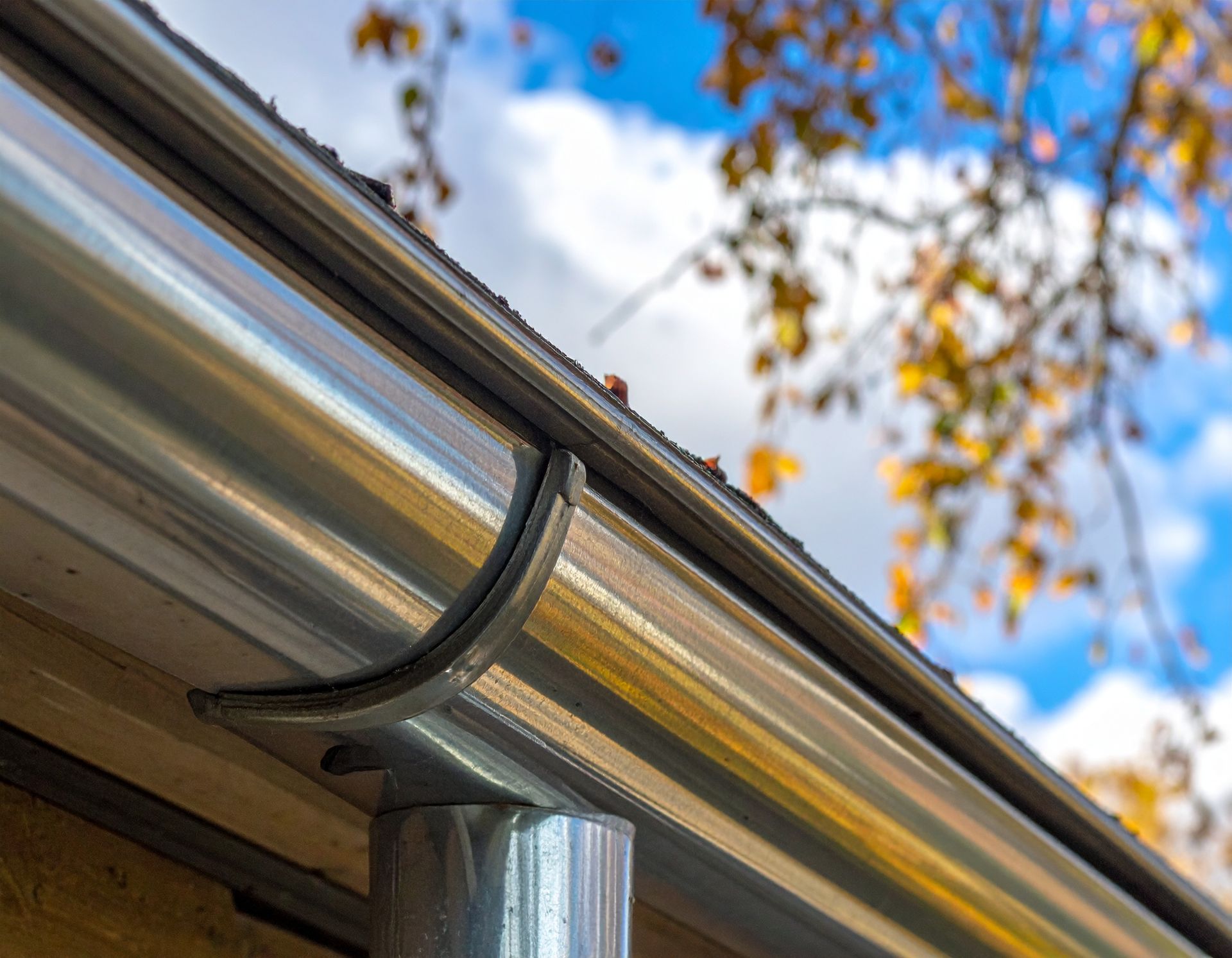 Gutter and downspout on a house roof with autumn tree branches and blue sky above