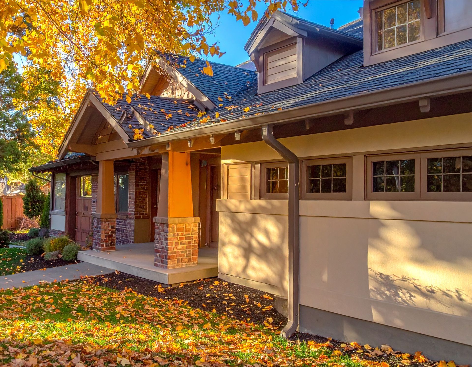 Sunlit suburban house with a front porch, beige garage, and autumn leaves on the lawn.