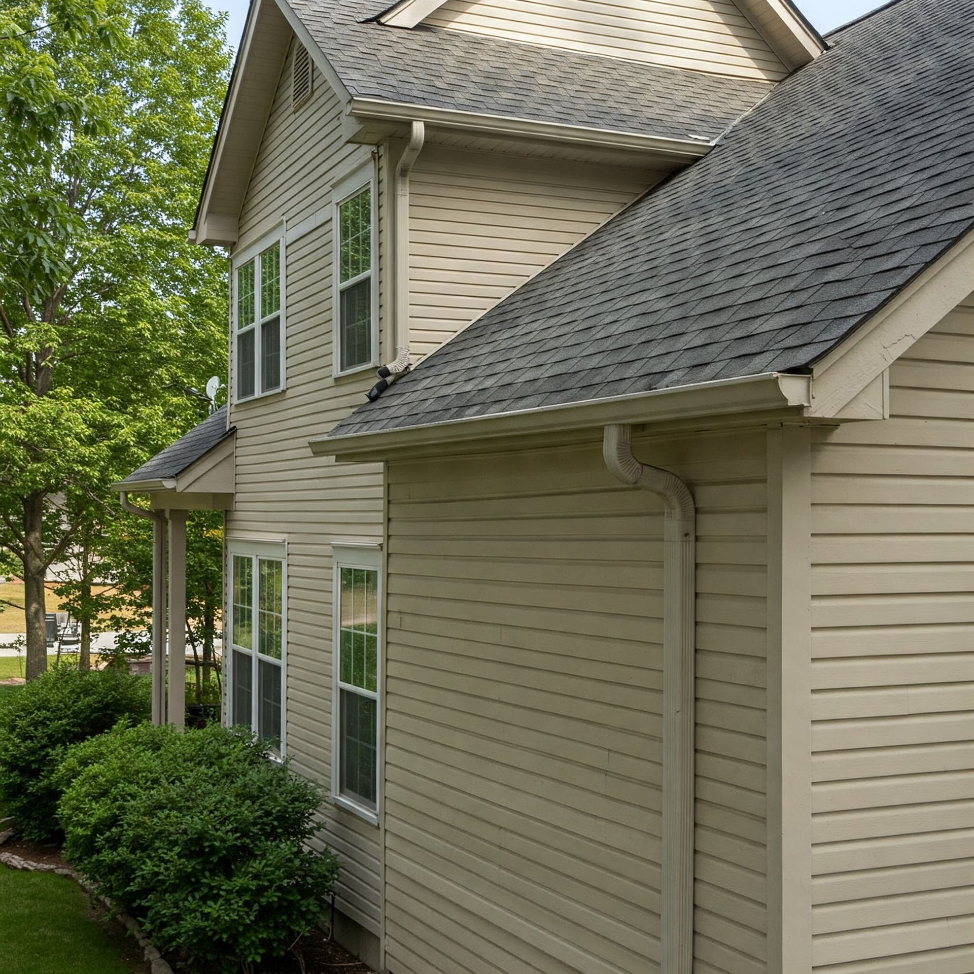 Beige house with gray roof and white trim, surrounded by green bushes and trees.