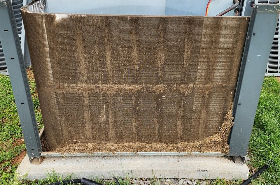 Rusty metal air conditioner condenser unit on a concrete pad outdoors, with a damaged brown mesh screen.