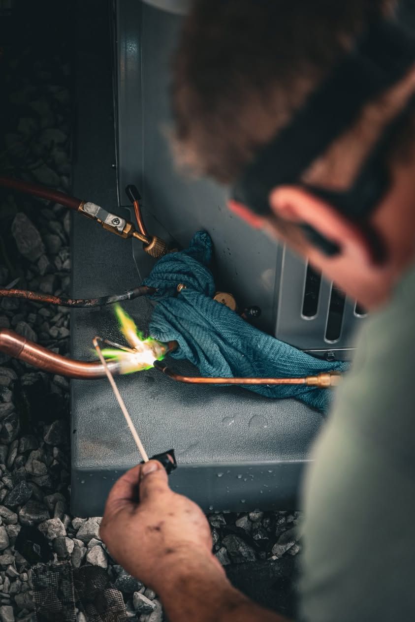 Technician wearing safety glasses brazes copper pipes on an outdoor HVAC unit using a torch and welding rod.