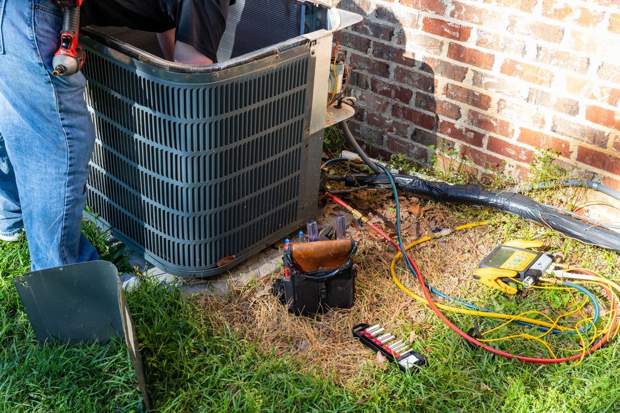 Outdoor HVAC unit being serviced with tools and wires beside a brick wall and grass.