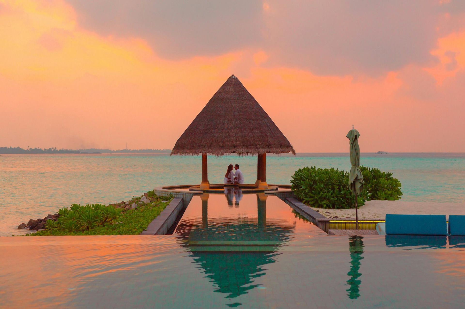 Couple in a thatched gazebo overlooking turquoise water at sunset; infinity pool in the foreground.