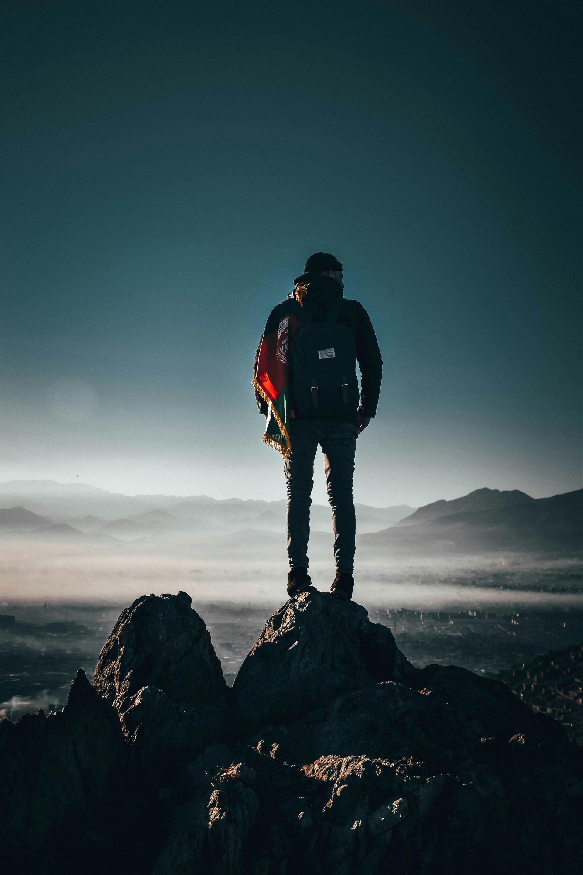 Person stands atop a rocky peak, holding a saxophone, overlooking a misty valley and mountains.