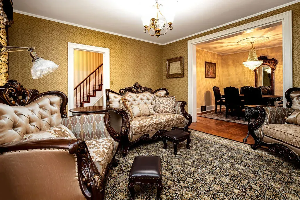 An ornate living room with gold patterned wallpaper, plush patterned furniture, and a view into a matching dining room.