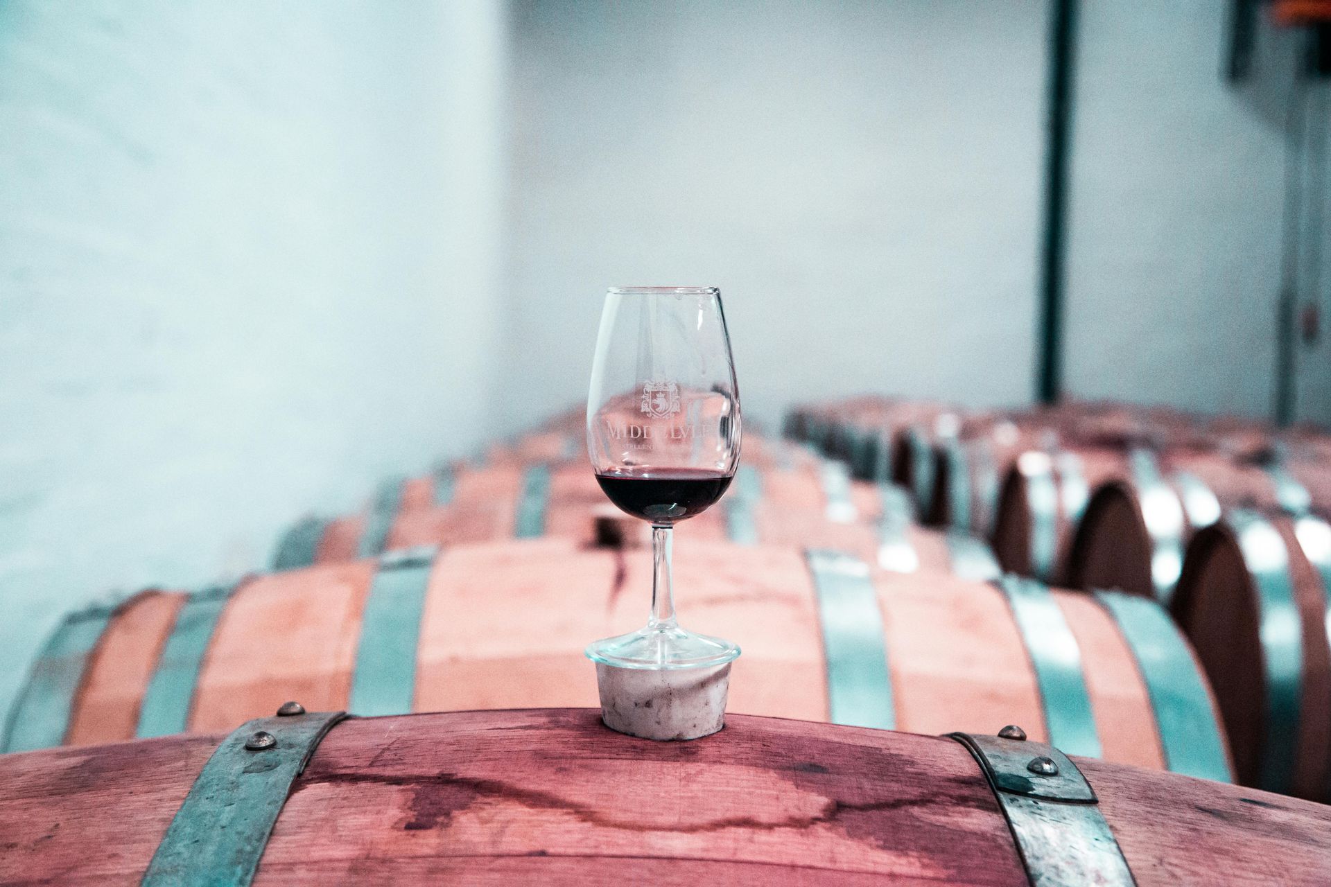 A wine glass filled with red wine sits on a wooden barrel in a cellar lined with rows of barrels.