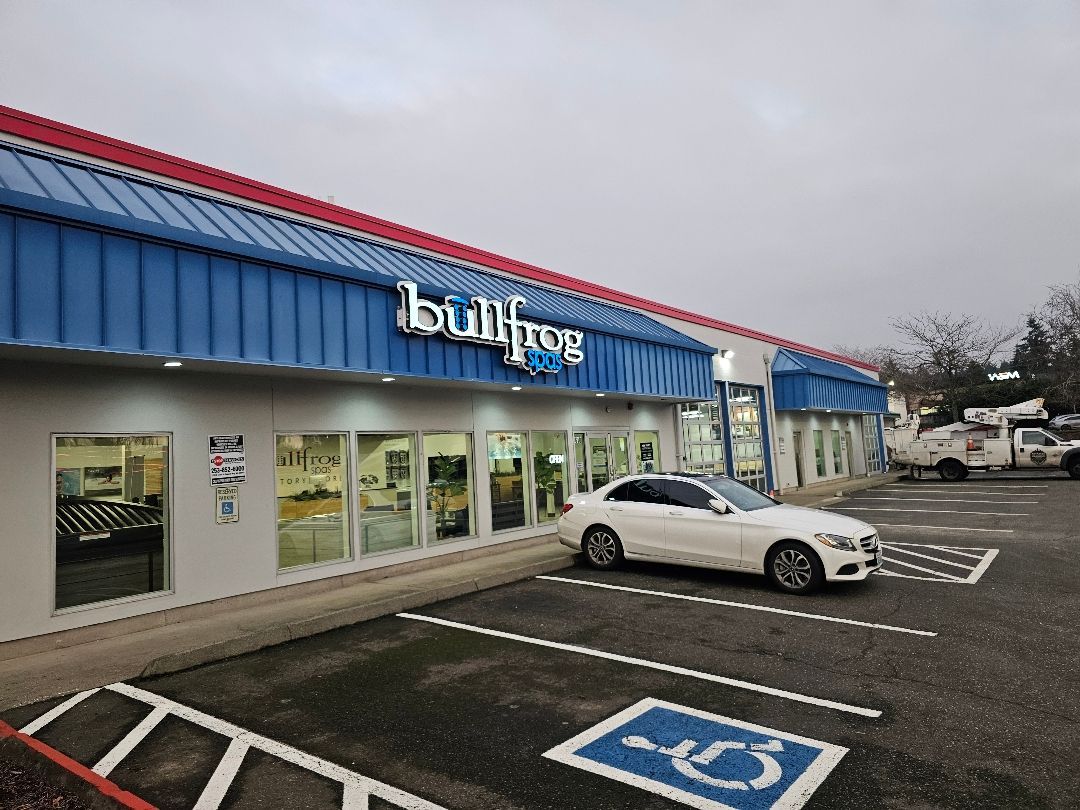 A Bullfrog Spas retail store with a blue metal roof, a white sedan parked out front, and a blue handicapped parking space.
