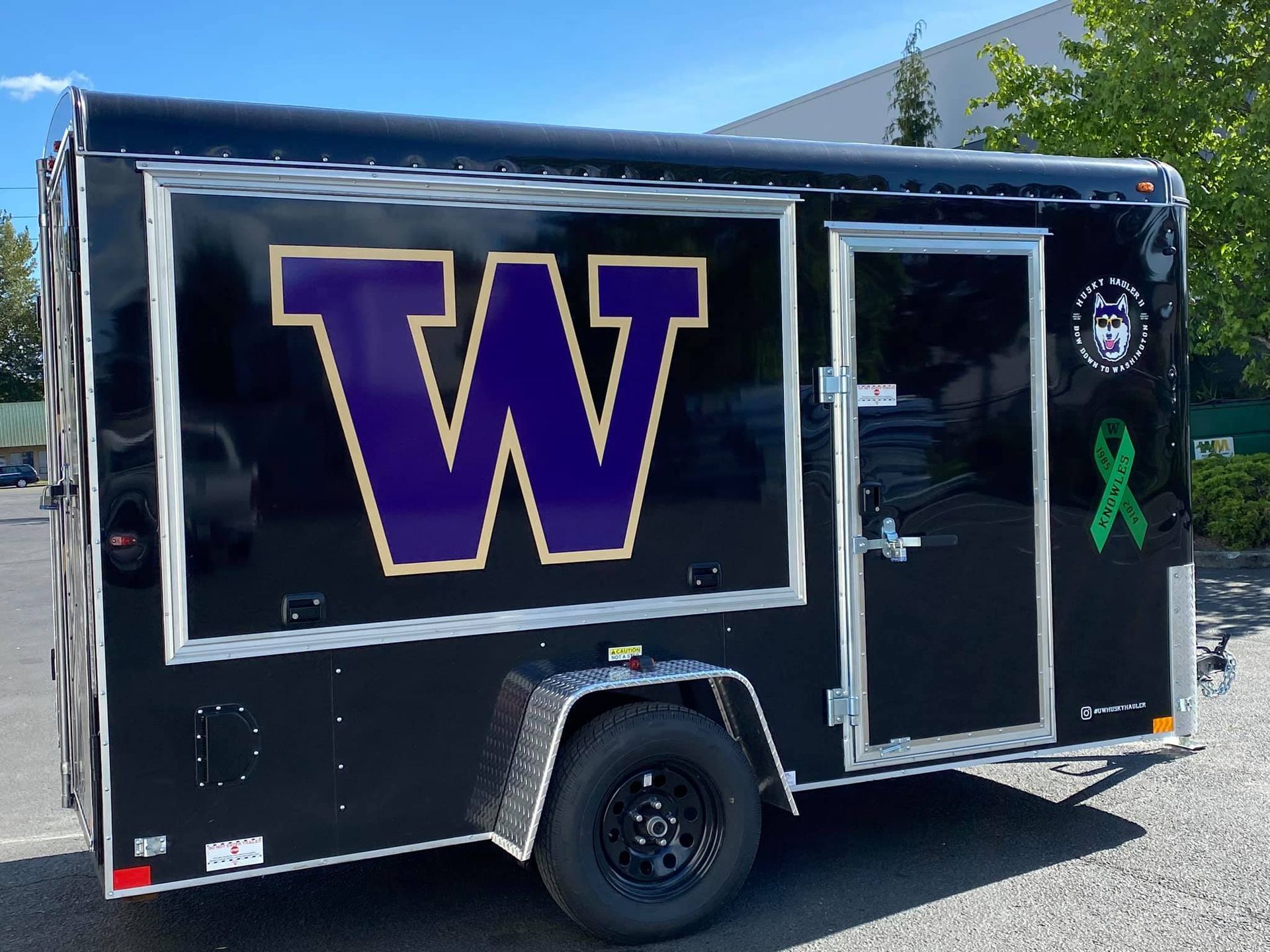A black trailer with a large purple and gold University of Washington 'W' logo, a husky emblem, and a green ribbon.