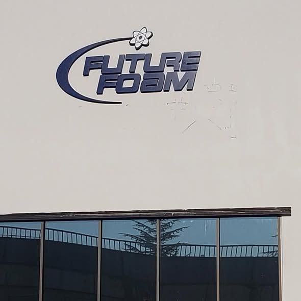The Future Foam logo in blue 3D lettering with an atom symbol above, mounted on a white exterior wall above windows.