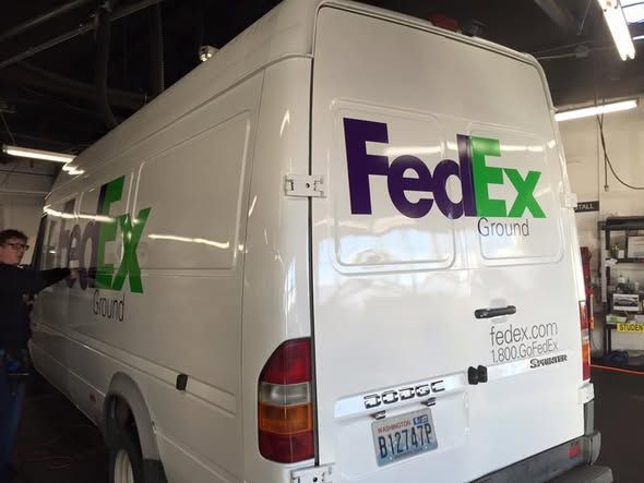 A white FedEx Ground cargo van parked indoors, showing a person applying a logo decal to the side panel.