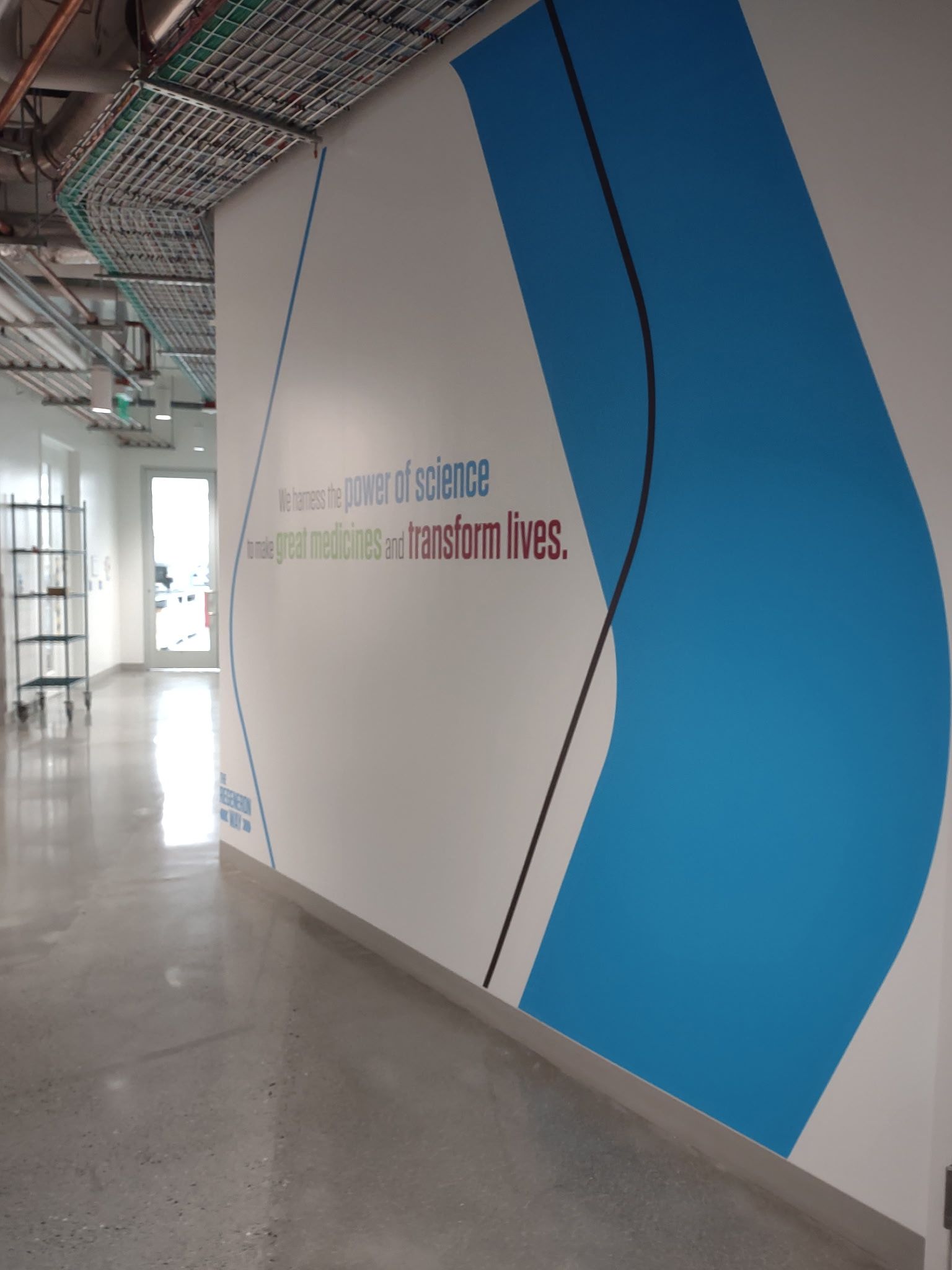 Indoor hallway with white wall and blue geometric mural, exposed ceiling, and polished concrete floor