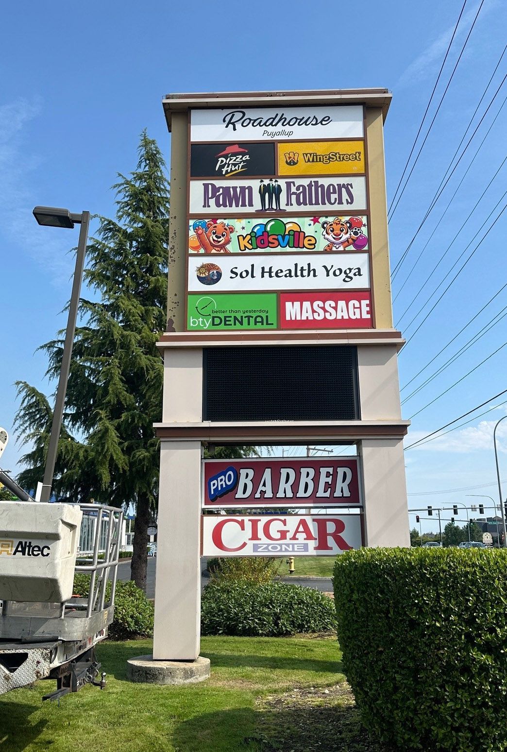 A tall, rectangular outdoor sign listing businesses including Pizza Hut, Pawn Fathers, and a cigar shop against a blue sky.