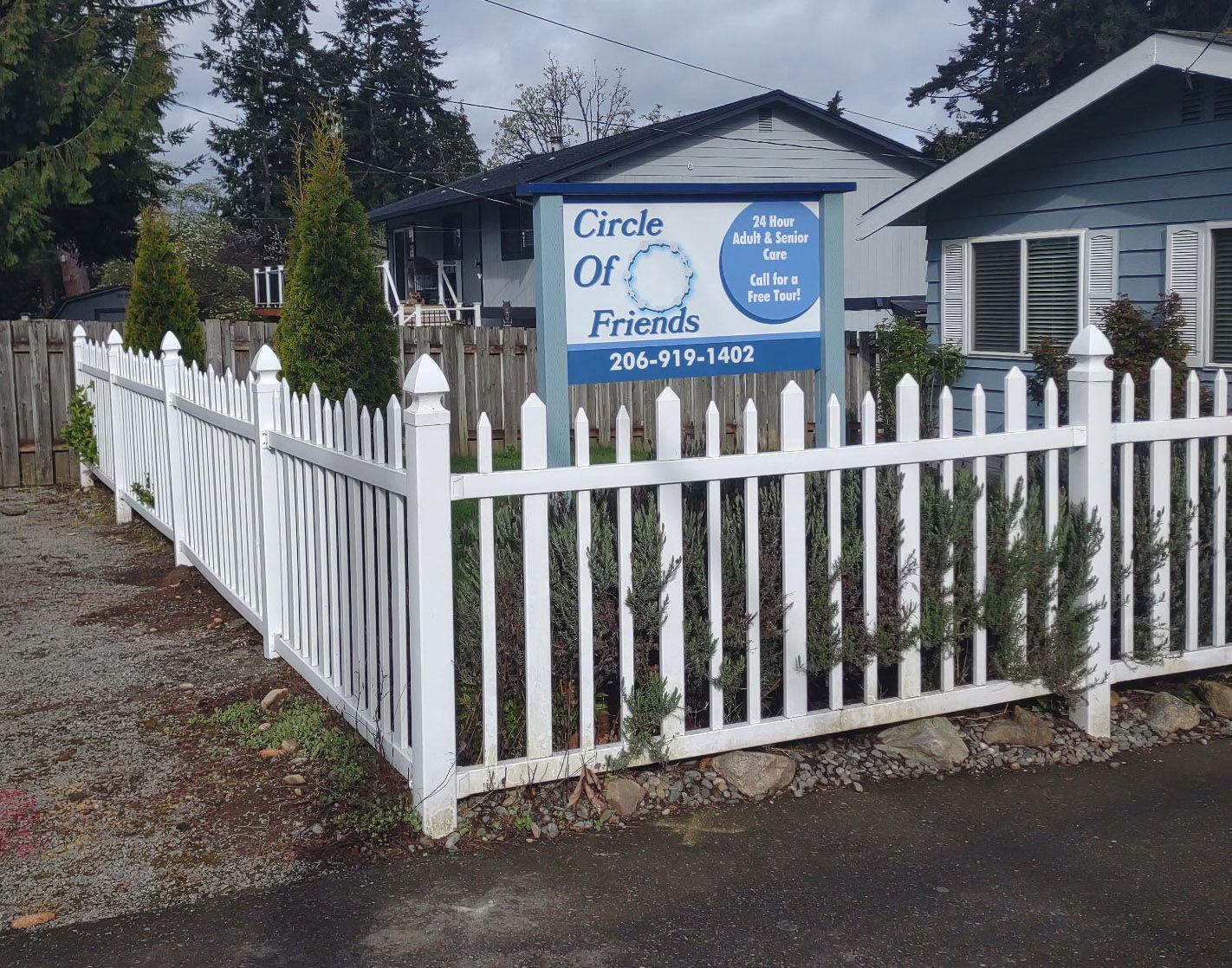 A white picket fence surrounds a yard with a sign for