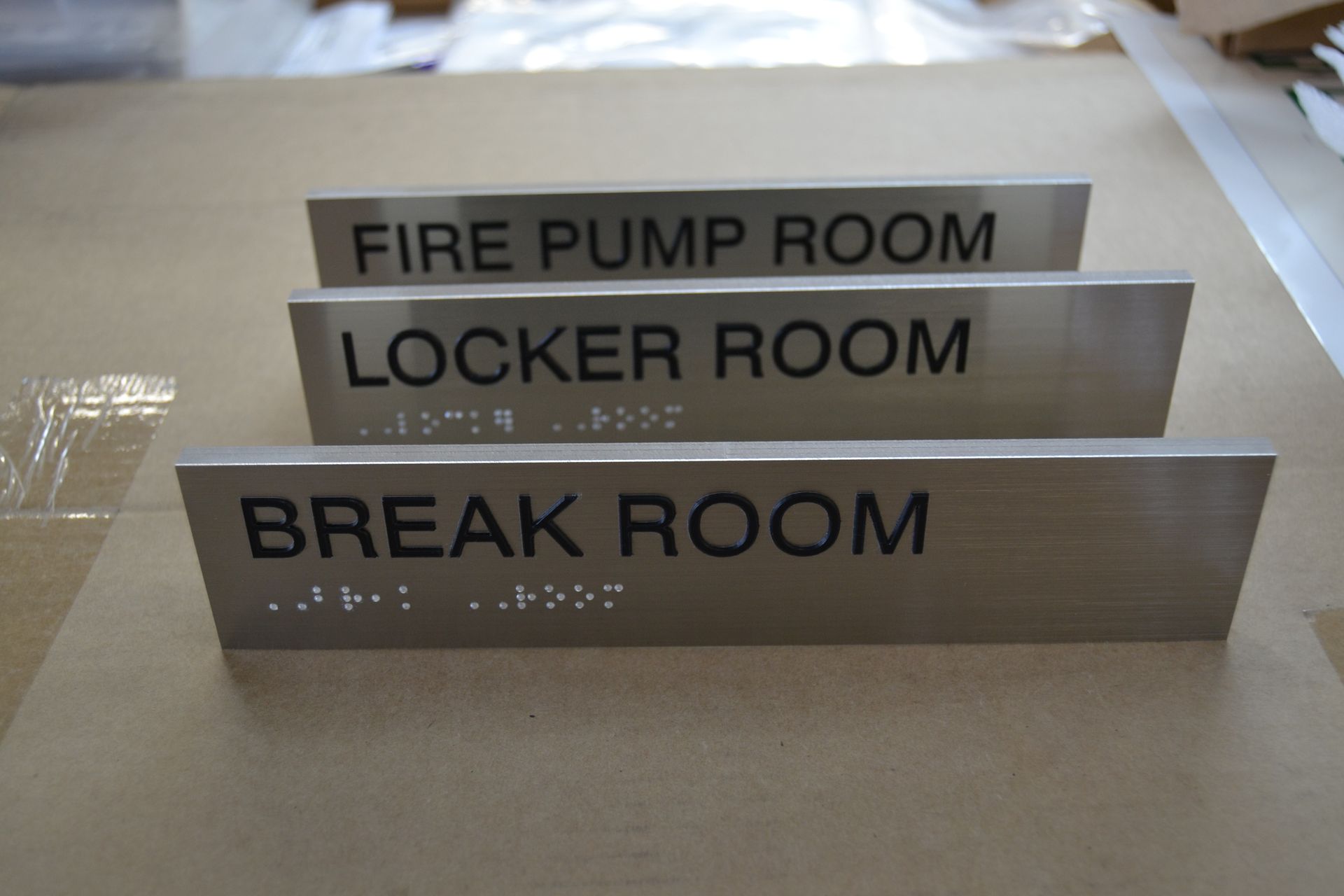Three brushed metal signs with black text and braille labeling rooms: FIRE PUMP ROOM, LOCKER ROOM, and BREAK ROOM.