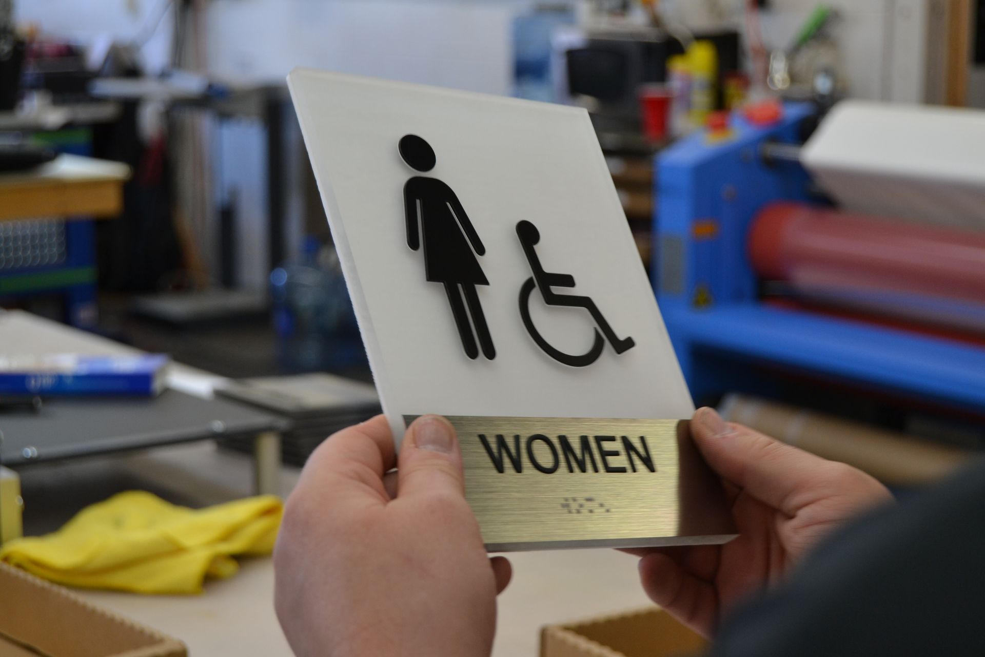 Hands hold a white, tactile ADA restroom sign featuring a black female figure and wheelchair icon, with 