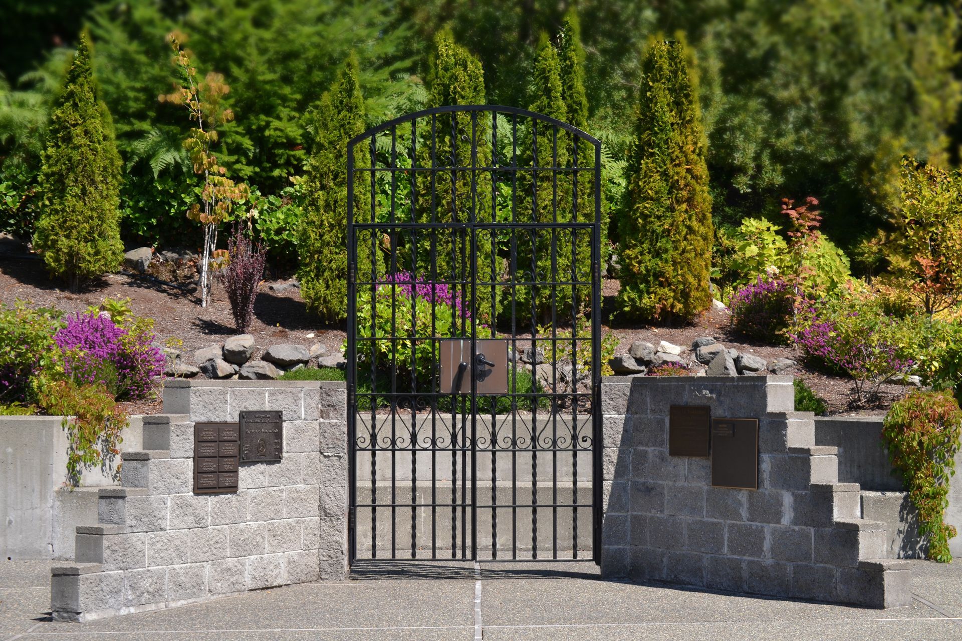 A tall black metal gate stands between two gray stone memorial walls featuring plaques, set against a garden background.