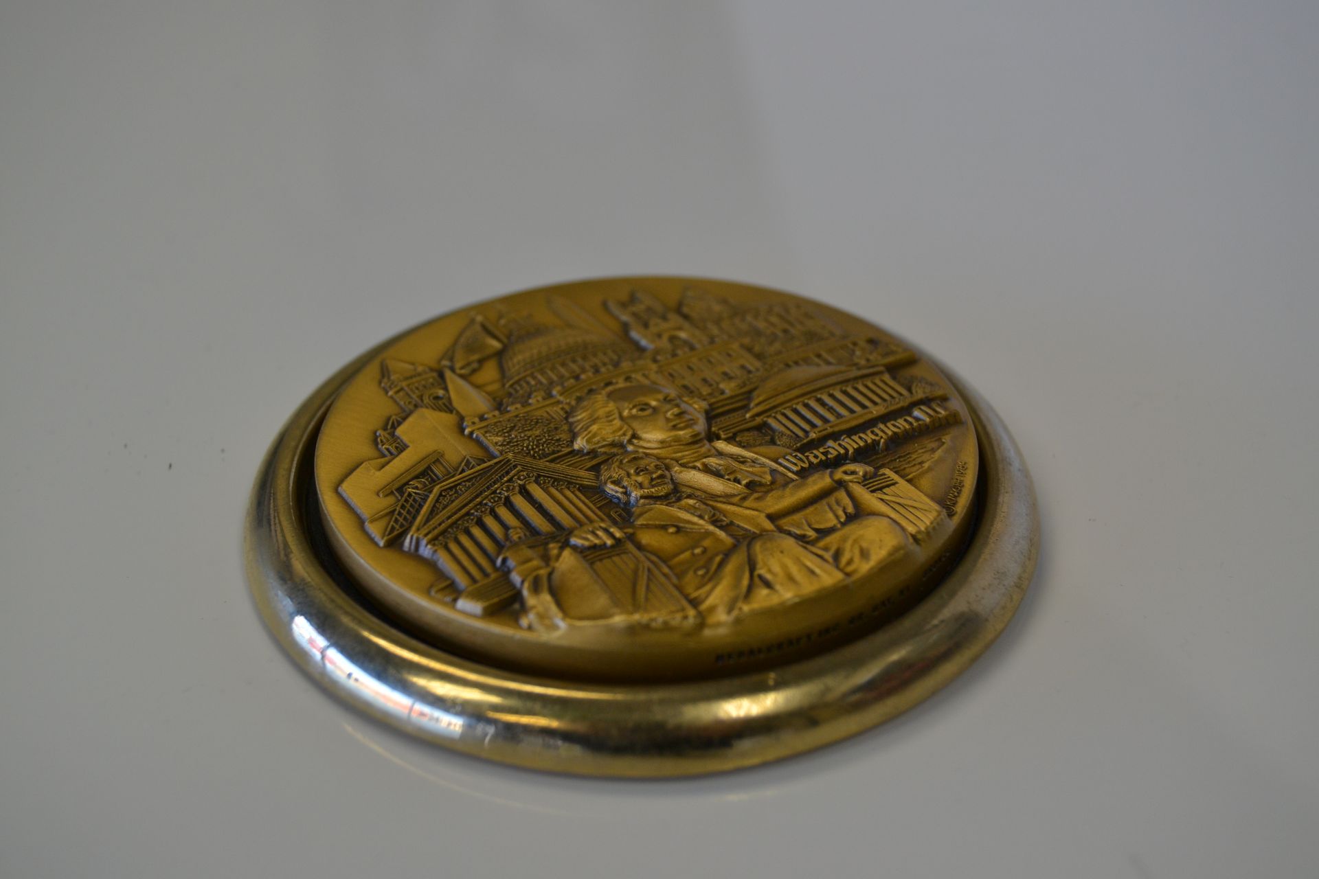 A round, gold-colored medallion featuring a detailed, raised relief of architectural landmarks, set in a silver-toned frame.