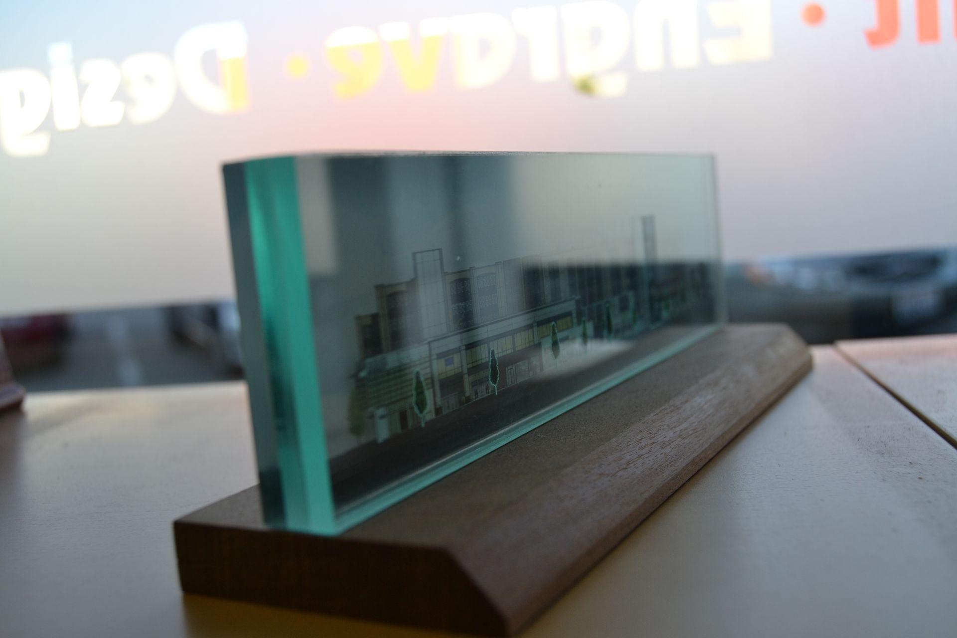 A rectangular glass block engraved with a building silhouette, mounted on a wooden base sitting on a desk.