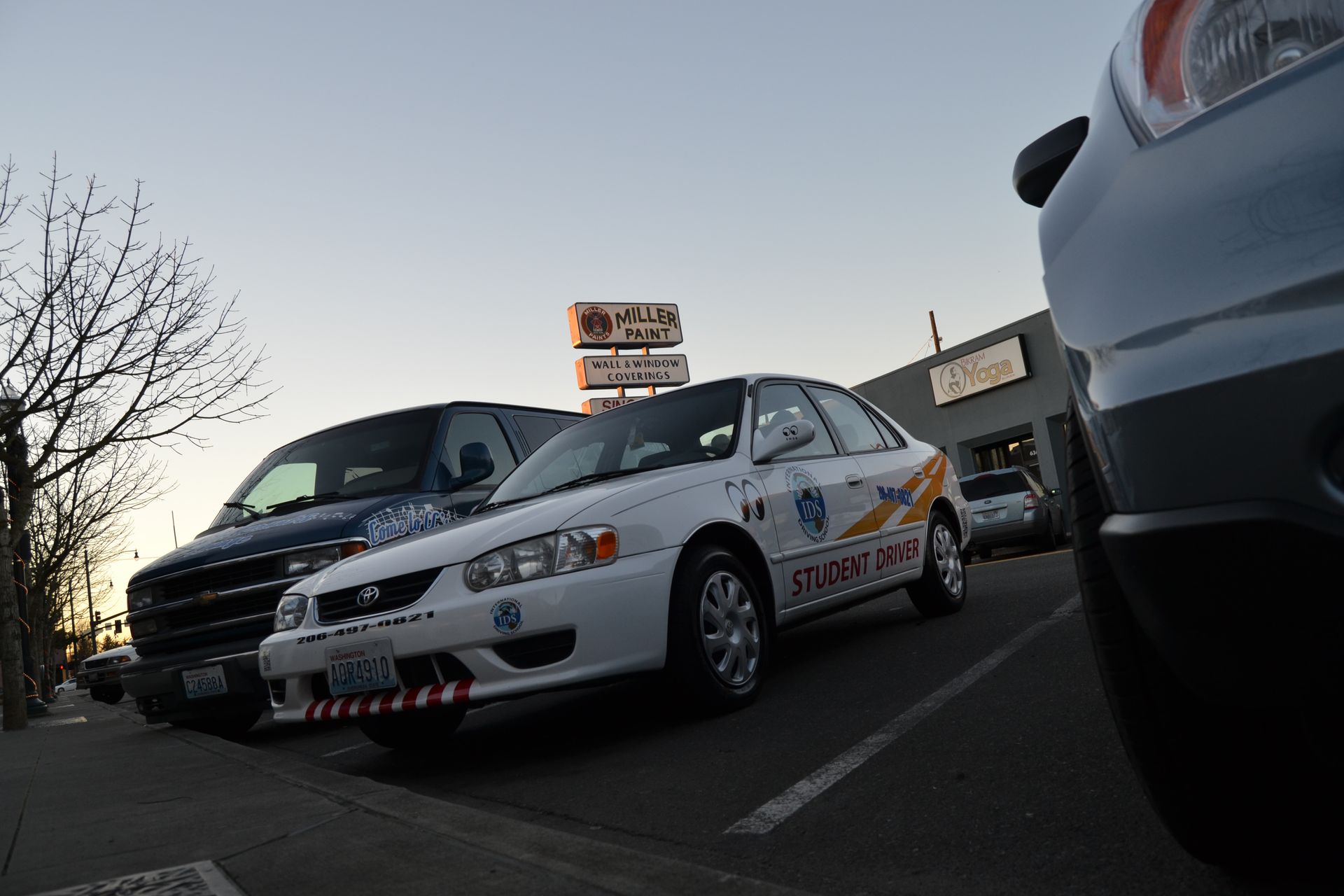 A white student driver car parked in a lot next to a black van at dusk, with a sign in the background.