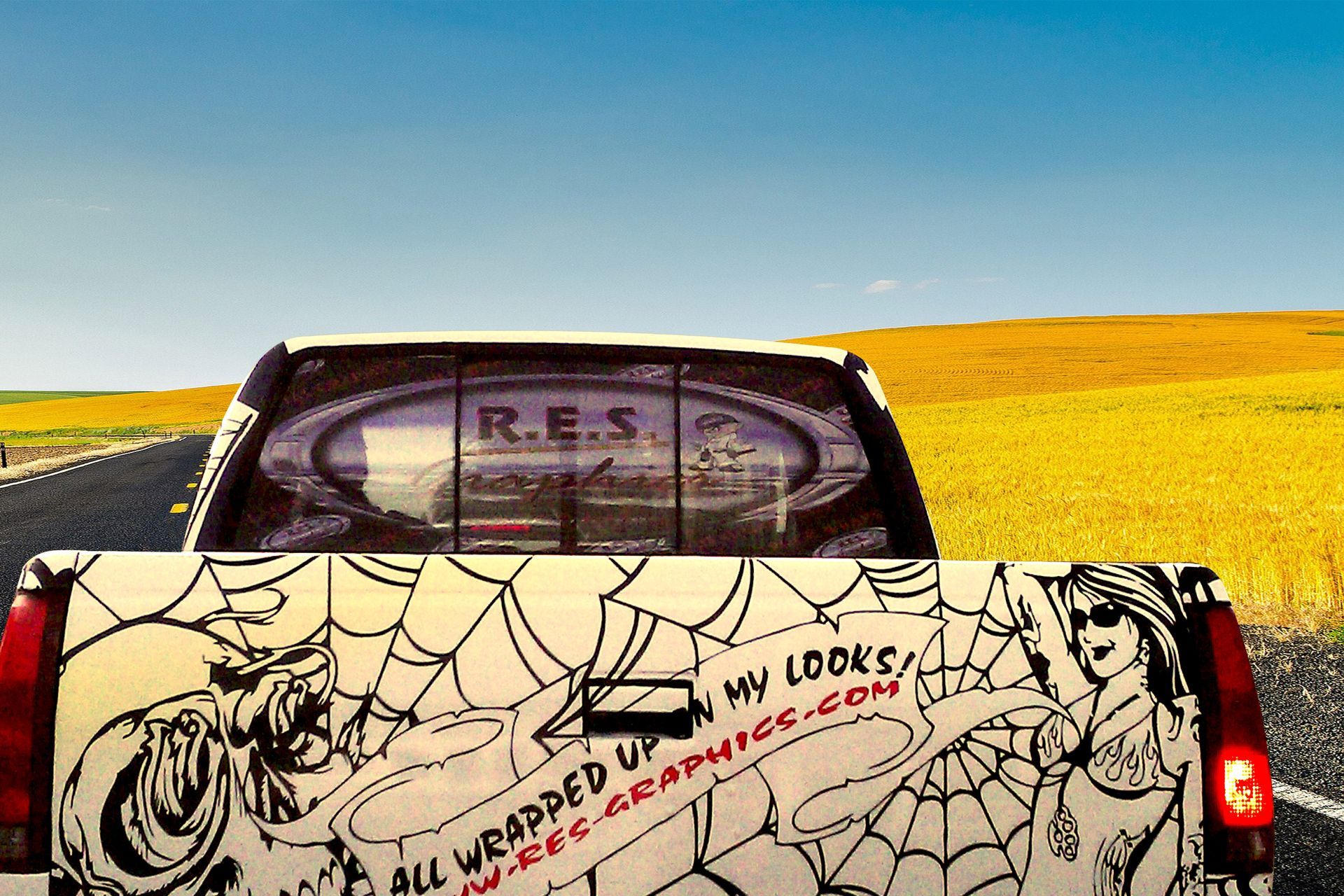 A pickup truck with a custom spiderweb-themed graphic on the tailgate and rear window, driving past a yellow field.