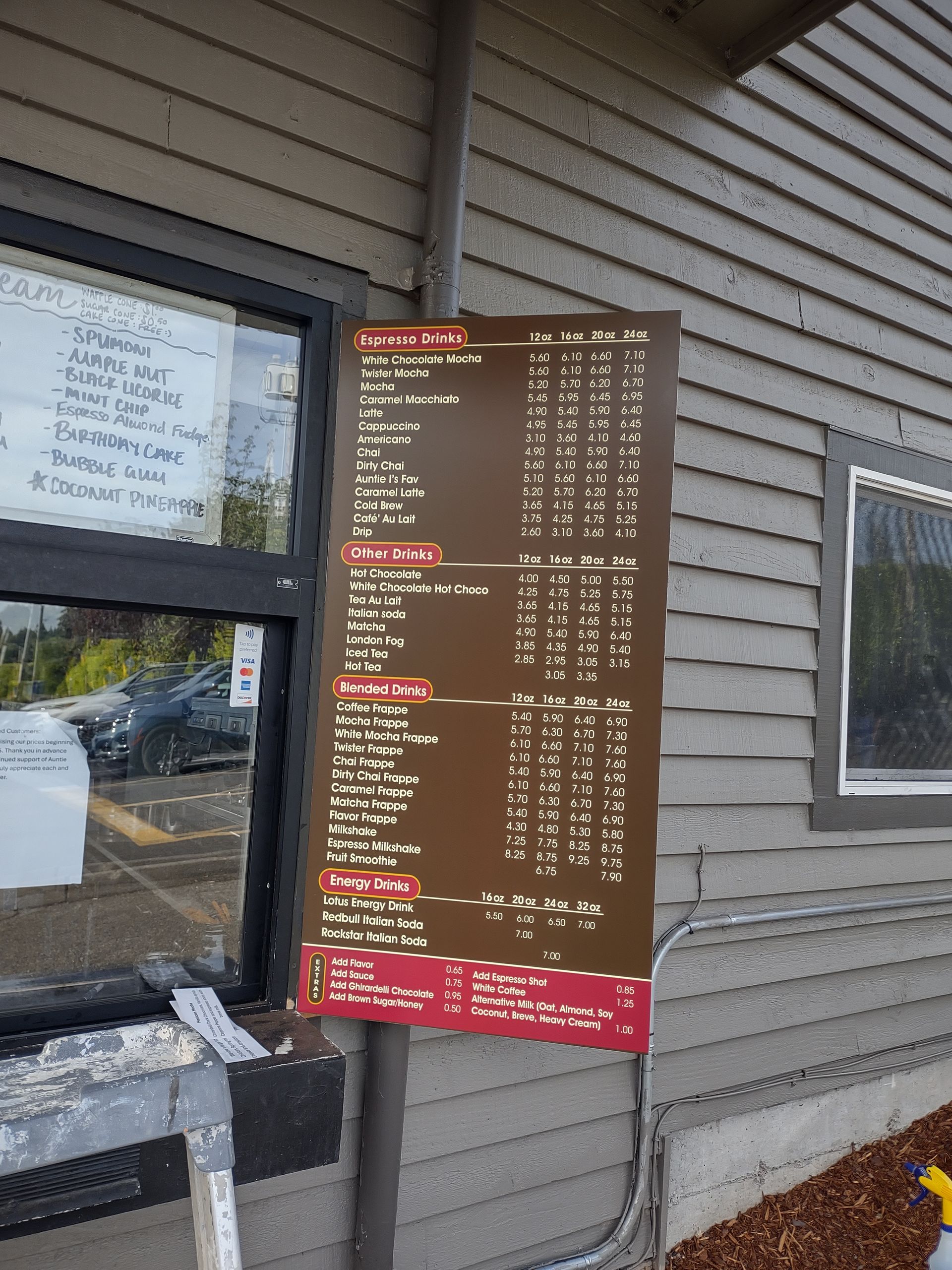 A brown menu board on the side of a building lists beverage and food options with prices in three columns.