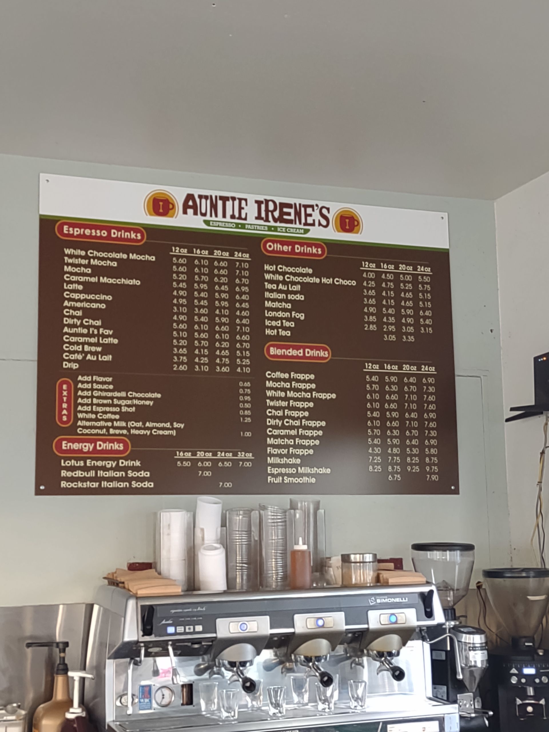 Auntie Irene’s coffee shop menu board lists various drink prices above a commercial espresso machine.