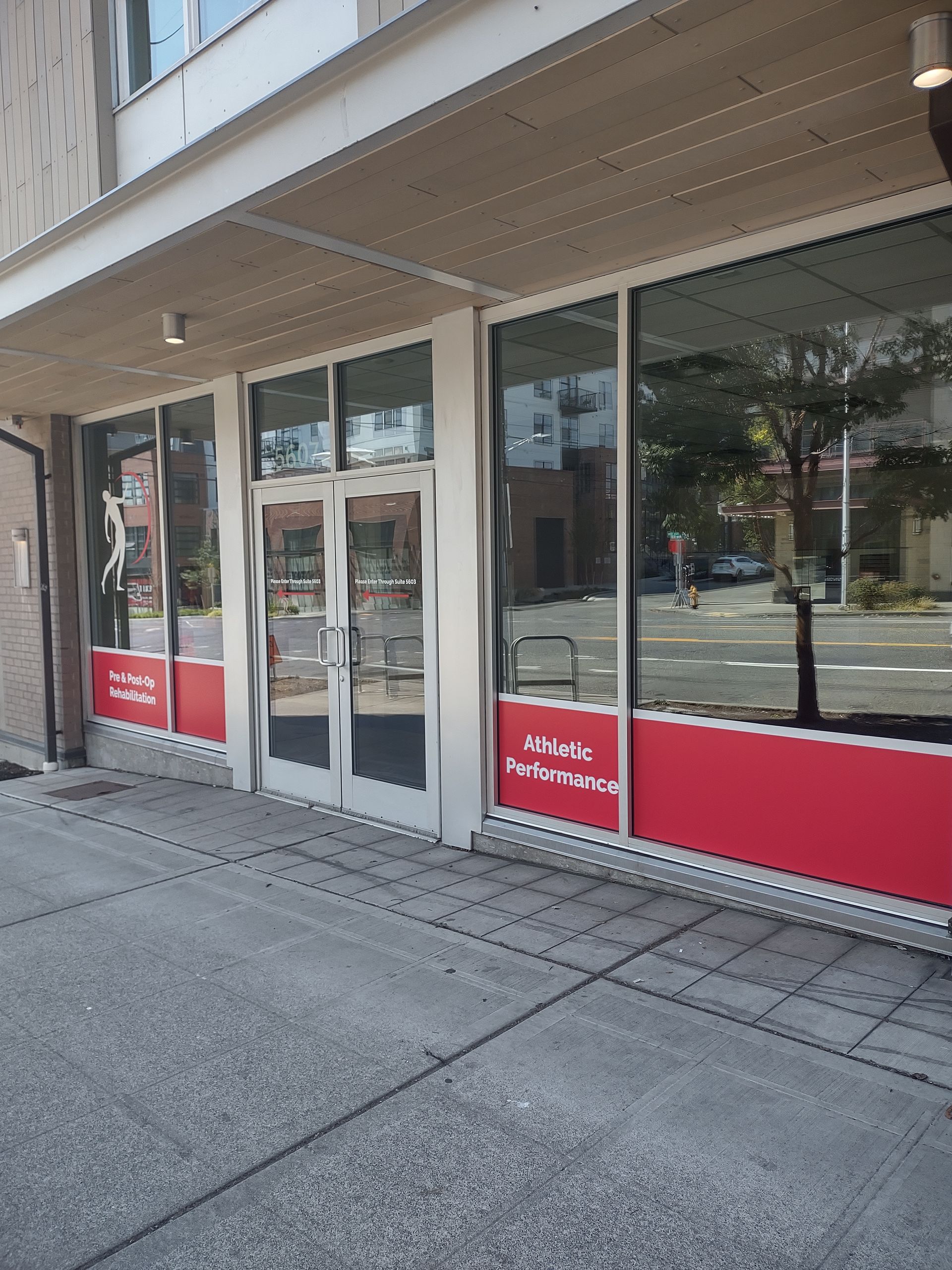 A store front with large glass windows featuring red signage panels, double glass entry doors, and a sidewalk entrance.