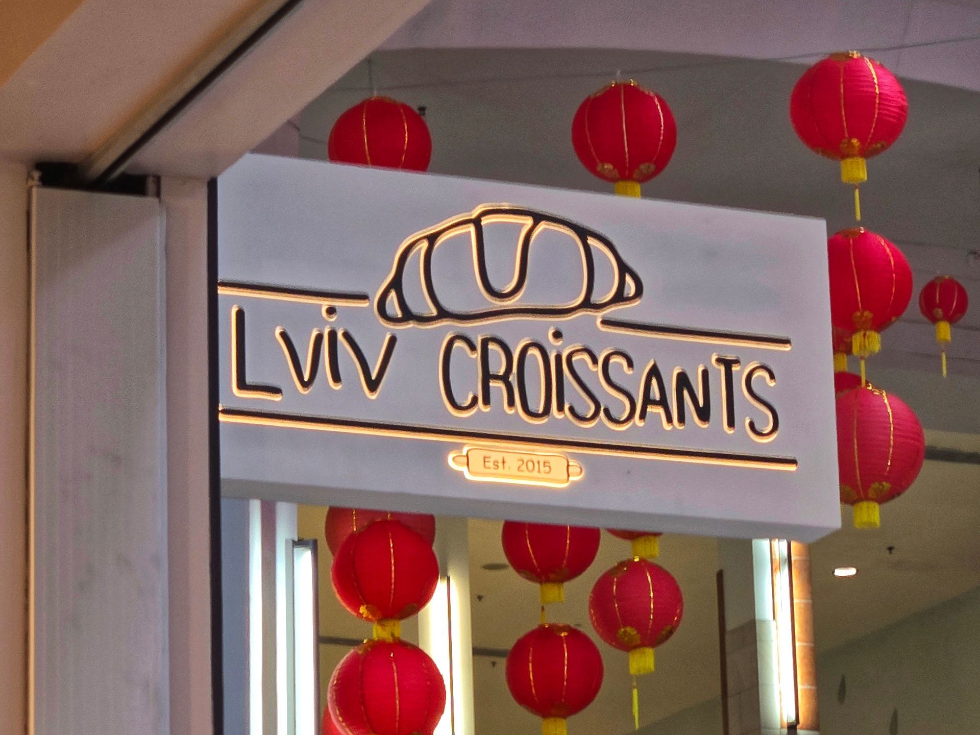 A backlit Lviv Croissants bakery sign hanging indoors, decorated with multiple red paper lanterns in the background.