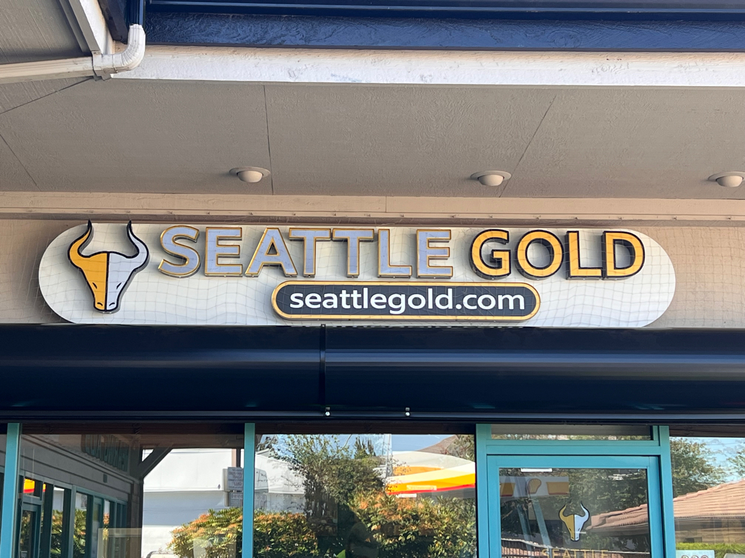 Storefront sign for Seattle Gold, featuring the business name and website on a beige oval plaque with a bull head logo.