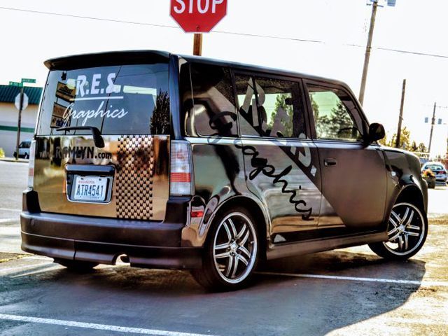 A dark-colored, custom-wrapped Scion xB parked in a lot, featuring