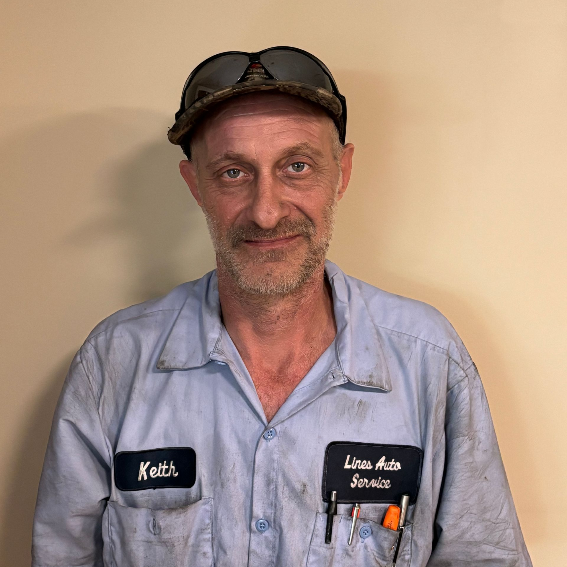 Smiling mechanic in a blue work shirt and cap, standing against a plain wall