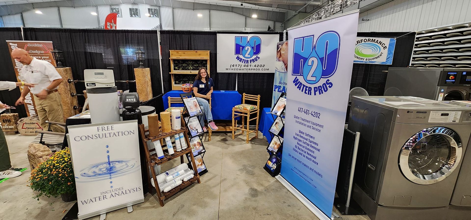 Booth display with stacked boxes, folding chairs, and a large washer/dryer in an exhibition hall