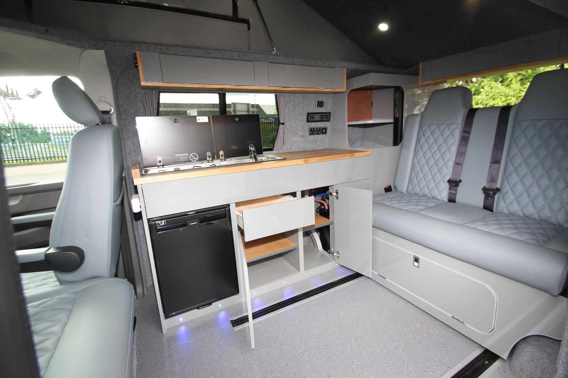 Interior of a van conversion with a compact kitchen, bench seating, and gray upholstery.