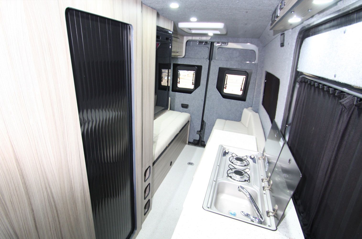 Narrow RV interior with bunk beds, white walls, black cabinets, and small windows.