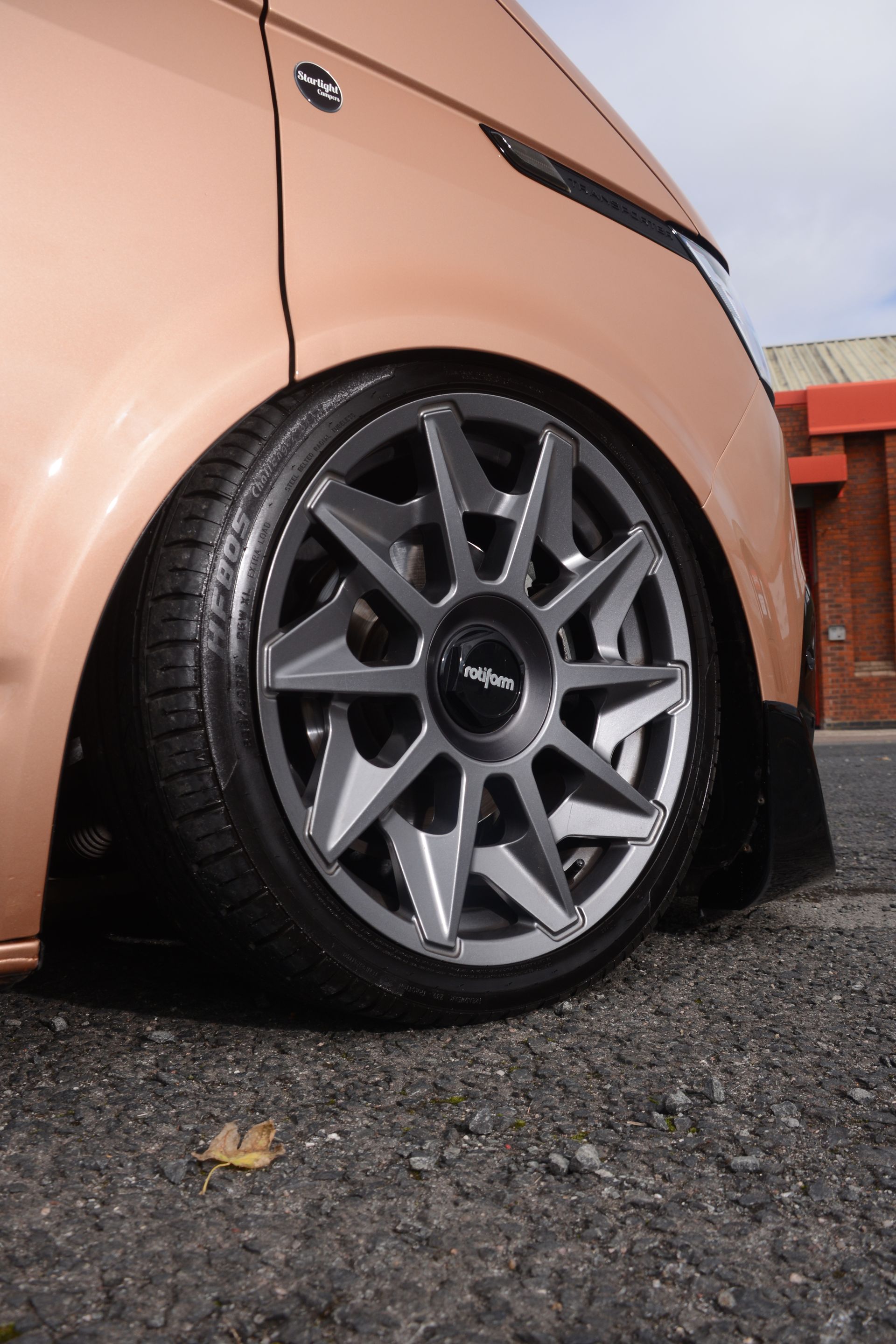 Close-up of a lowered peach car with a gray alloy wheel parked on wet gravel.