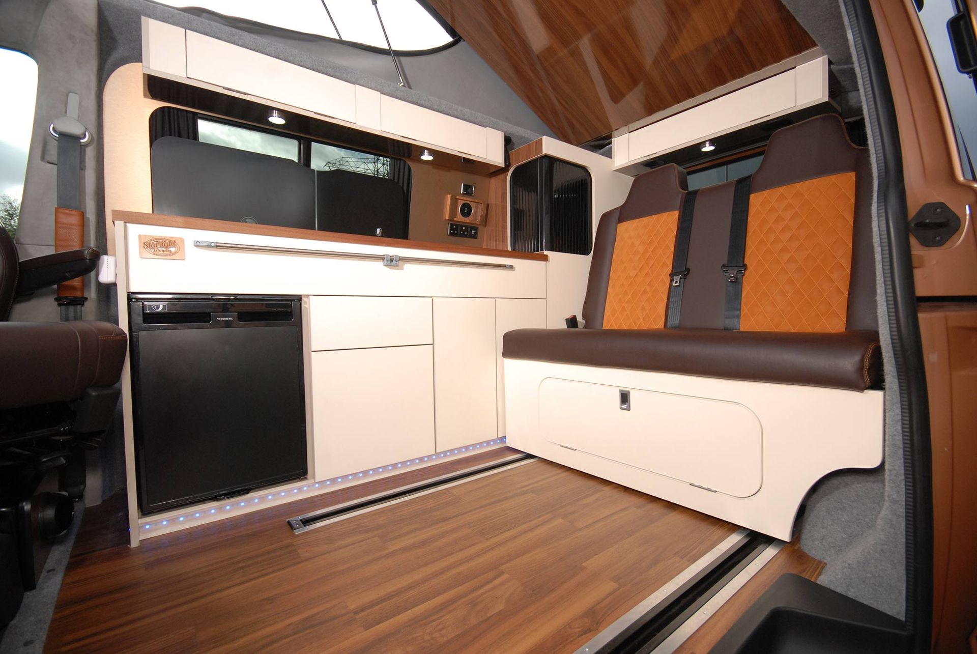 Compact camper van interior with wood flooring, bench seat, and kitchenette