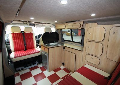 Compact camper van interior with red-and-white seats, wood cabinets, and checkered floor.