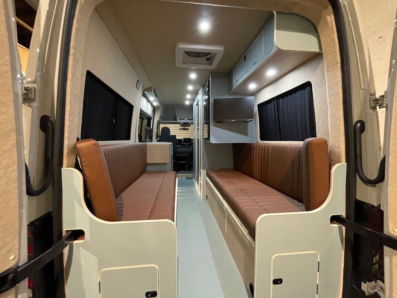 Empty camper van interior with brown bench seats, white cabinets, and black curtains