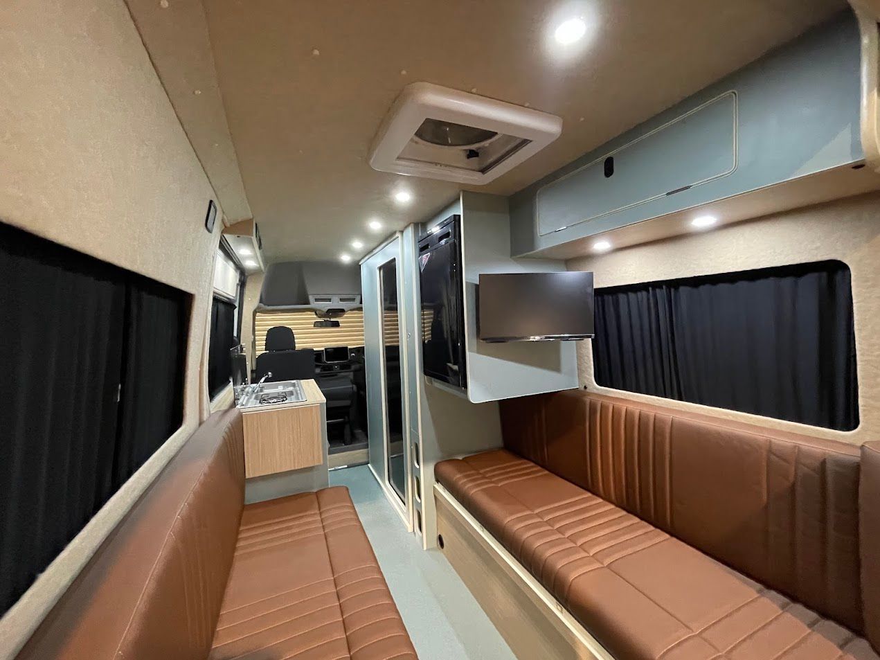 Narrow train sleeper cabin with brown benches, blue storage, curtained windows, and overhead lights.
