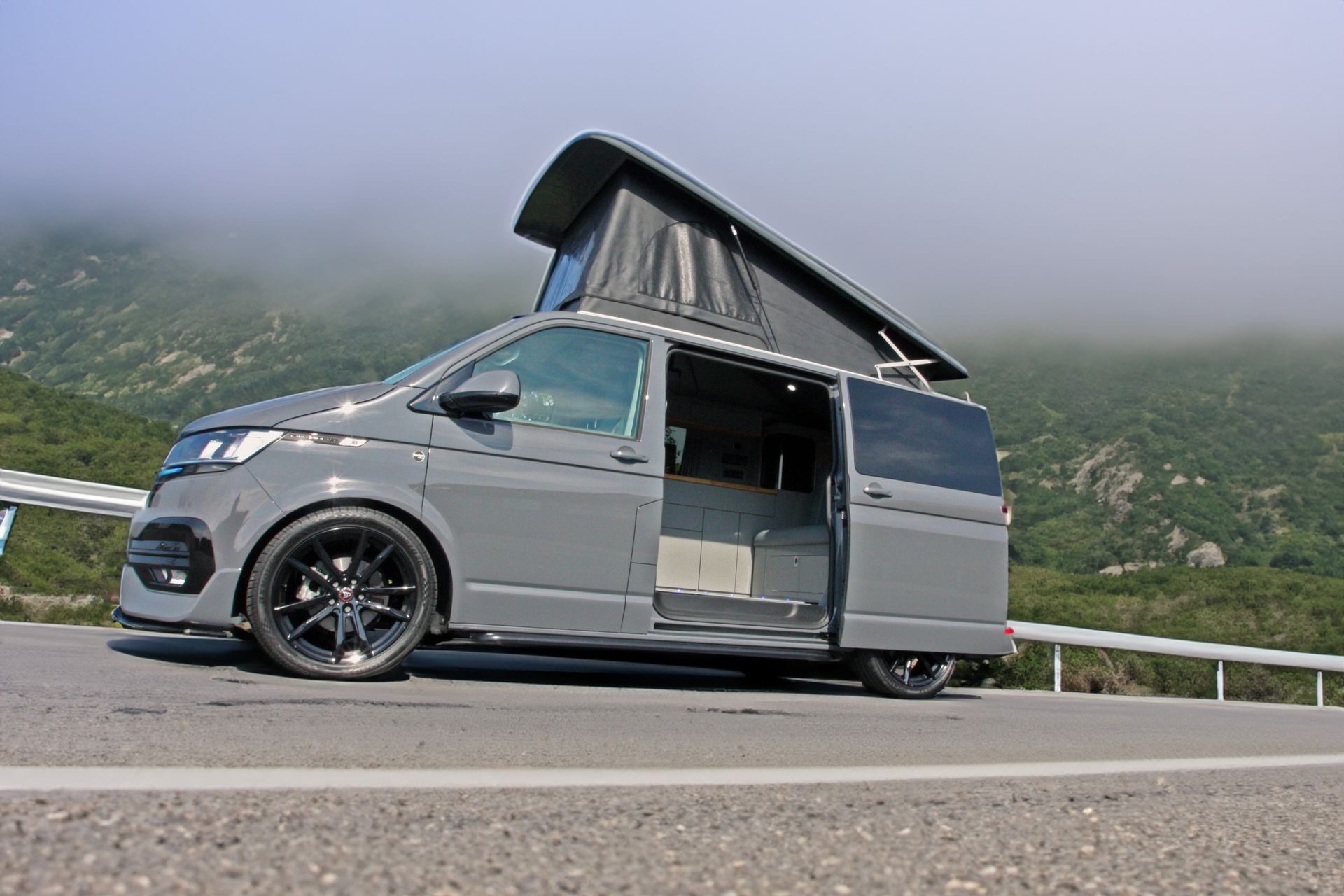 Gray camper van with pop-up roof parked on a mountain road with misty hills behind