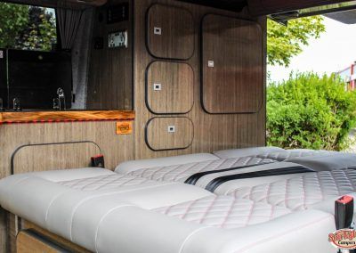 Inflatable mattress in a wood-paneled camper van with open side door and greenery outside
