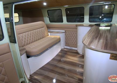 Compact camper interior with tan bench seating, wood floor, and small round table.