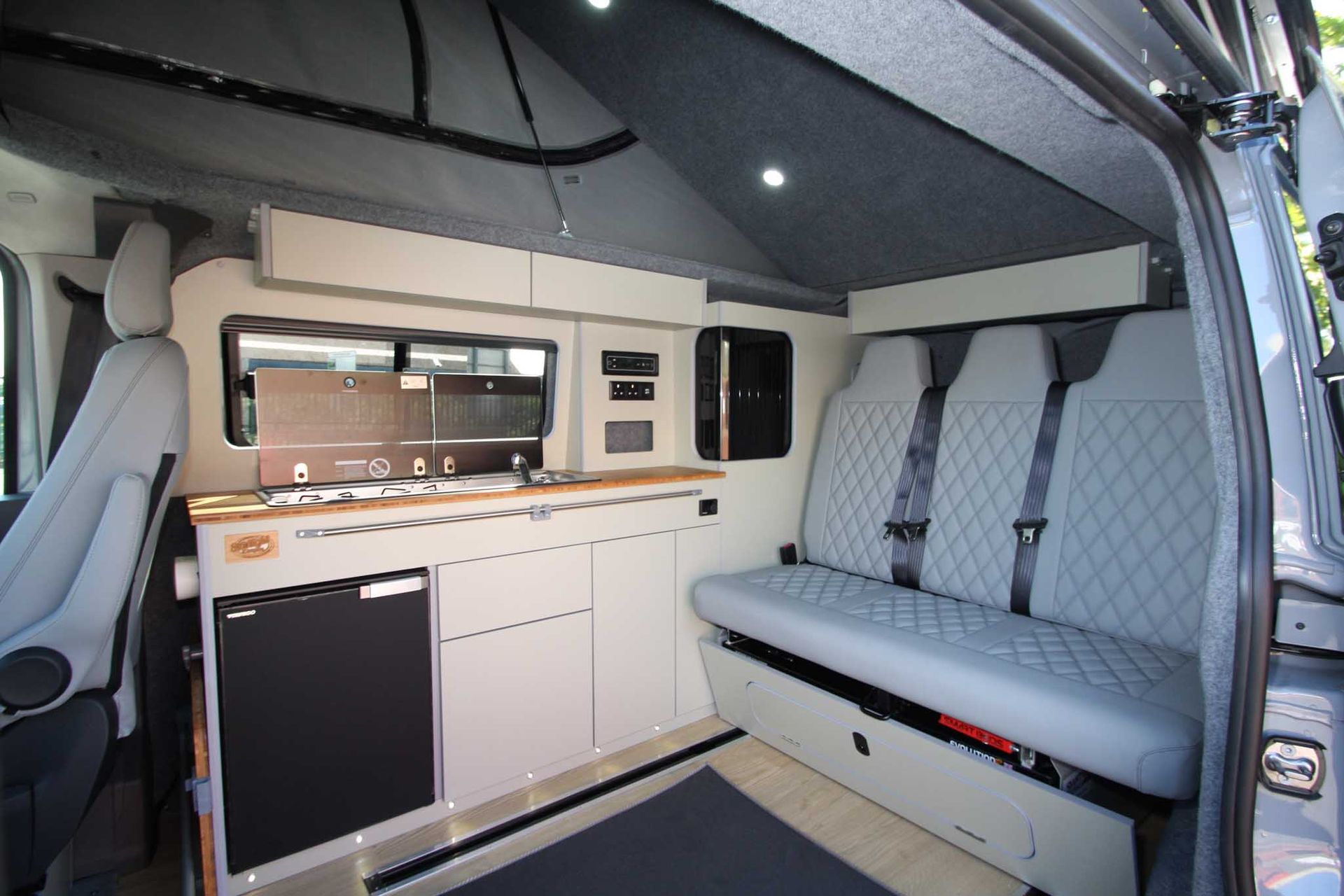 Compact camper van interior with white cabinets, bench seat, and quilted gray upholstery