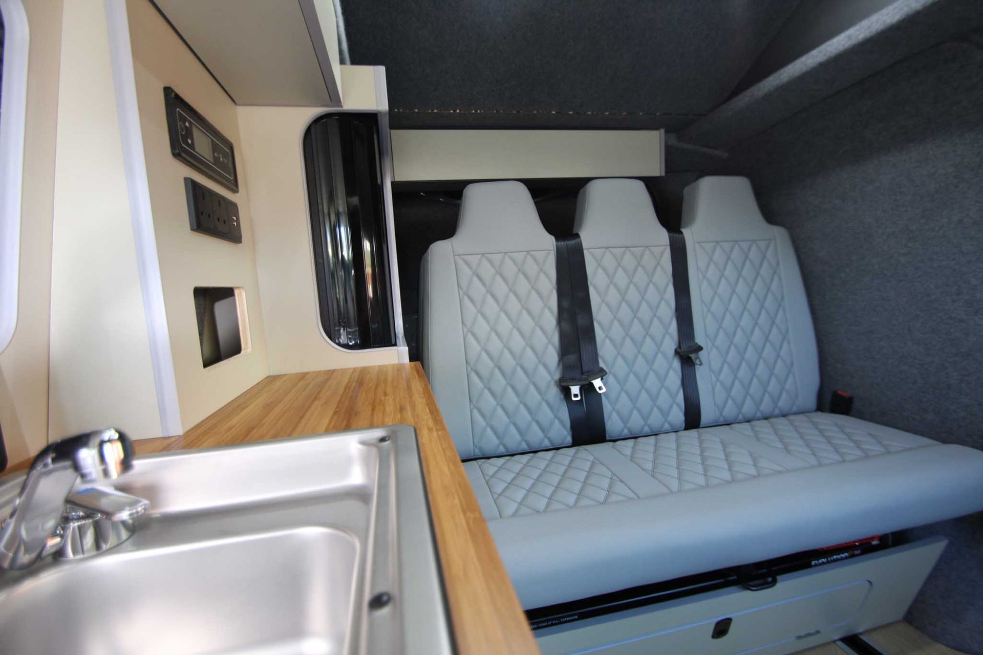 Compact camper van interior with sink, wood countertop, and gray quilted bench seats