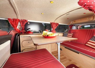 Red-upholstered camper van interior with table, bench seats, and curtains.