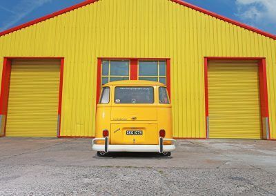 Yellow vintage van parked before bright yellow warehouse doors with red trim.
