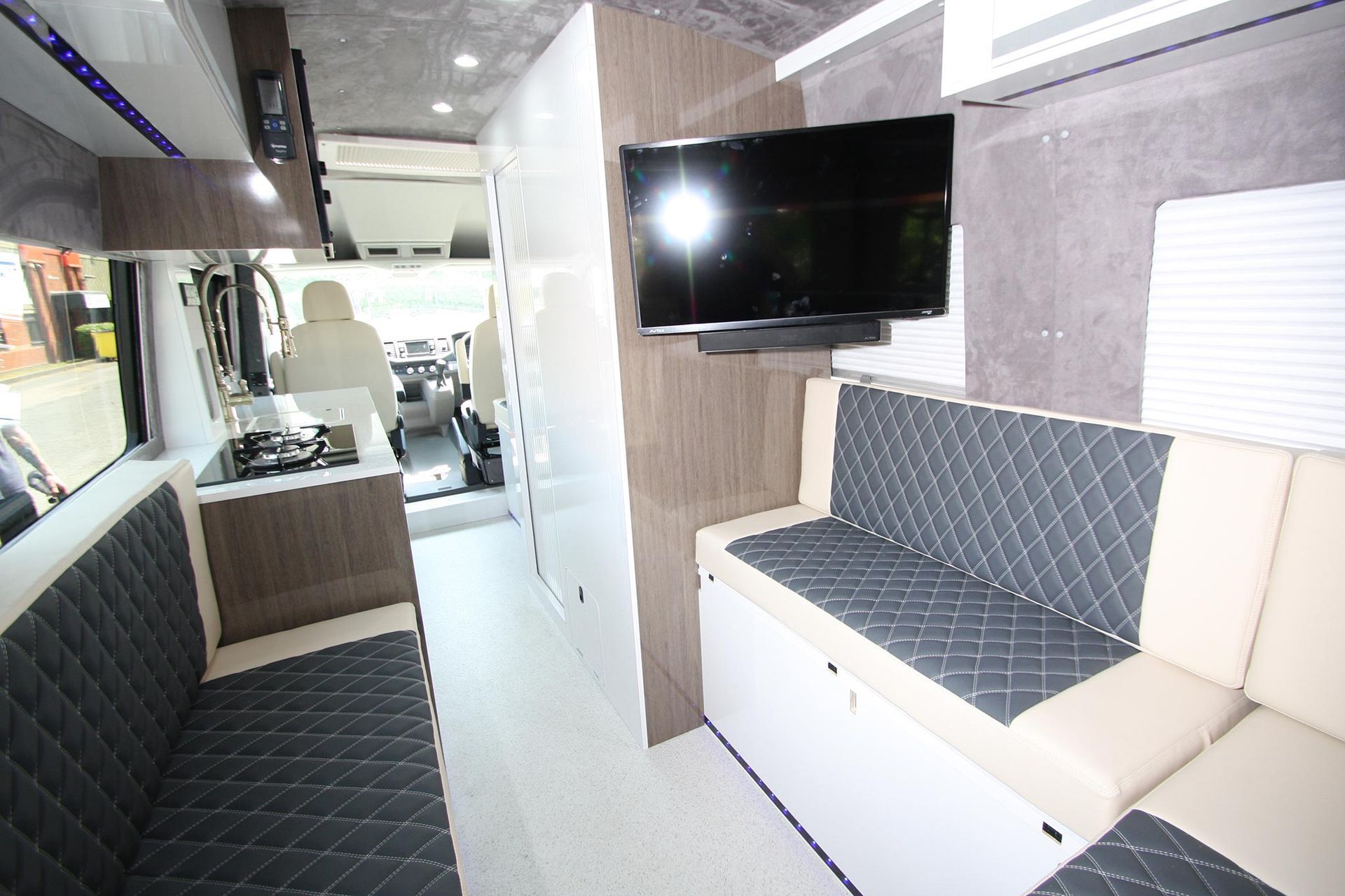 Bright camper van interior with quilted bench seating, wall-mounted TV, and a front cabin view.