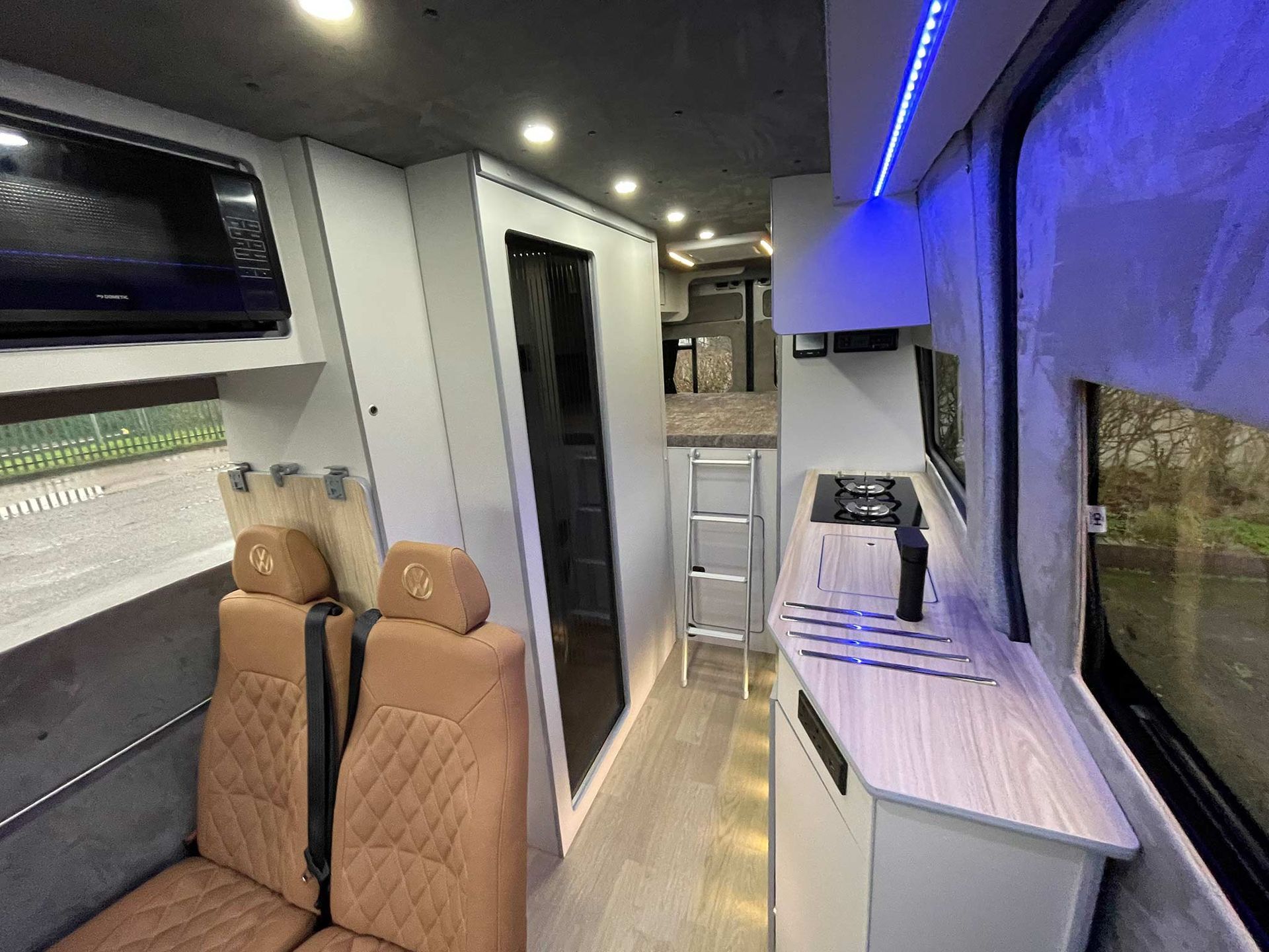 Modern RV interior with tan seats, white counters, blue LED lighting, and a side window